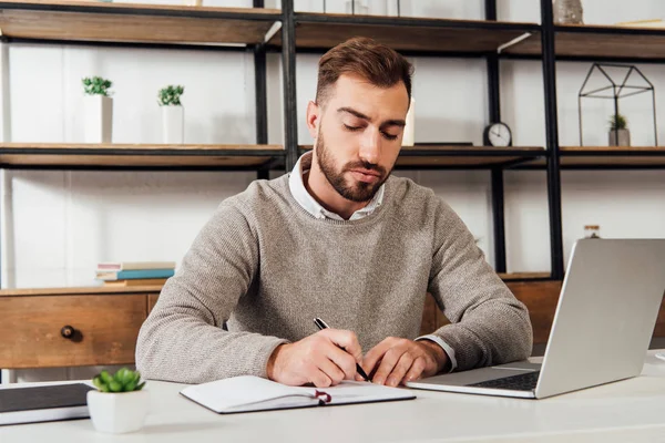 Mann schreibt auf Notizbuch neben Laptop auf Tisch — Stockfoto