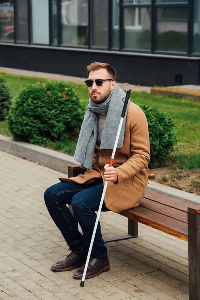 Aveugle en manteau tenant un bâton de marche assis sur un banc — Photo de stock