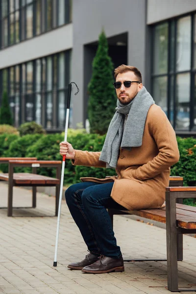 Hombre ciego en gafas de sol sosteniendo bastón mientras está sentado en el banco - foto de stock