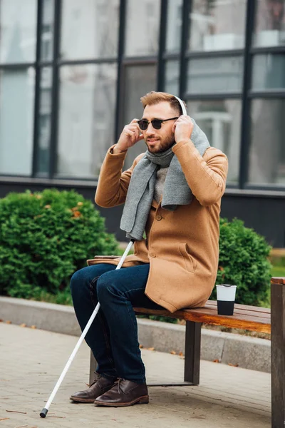 Hombre ciego usando auriculares mientras está sentado en el banco - foto de stock
