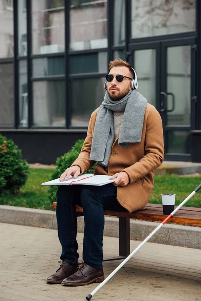 Hombre ciego en auriculares libro de lectura en el banco en el parque - foto de stock