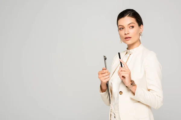 Beautiful pensive businesswoman in white suit holding clipboard and pen, isolated on grey — Stock Photo