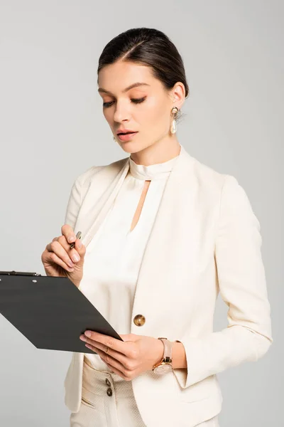 Hermosa mujer de negocios en traje blanco escrito en portapapeles, aislado en gris - foto de stock