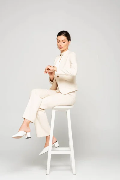 Elegante Geschäftsfrau im weißen Anzug schaut auf die Uhr, während sie auf einem Schemel sitzt, auf grau — Stockfoto