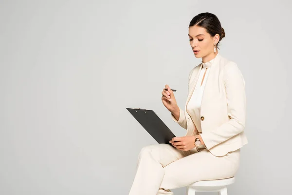 Елегантна приваблива бізнес-леді в білому костюмі, сидячи на табуреті з ручкою і буфером, ізольована на сірому — стокове фото