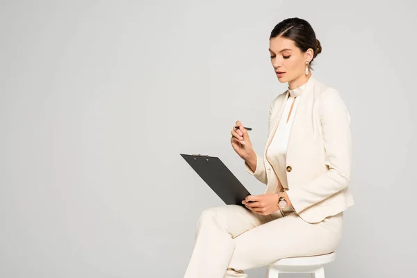 Elegante mujer de negocios atractiva en traje blanco sentado en el taburete con la pluma y el portapapeles, aislado en gris - foto de stock