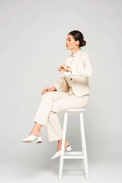 Elegant businesswoman in white suit sitting on stool, on grey — Stock Photo