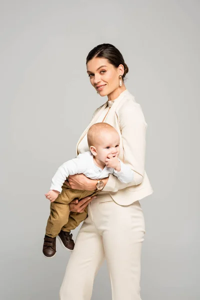 Bela mulher de negócios alegre segurando pequeno bebê a mãos, isolado em cinza — Fotografia de Stock