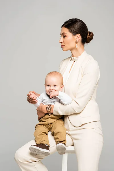 Beautiful businesswoman with little happy baby on hands, isolated on grey — Stock Photo