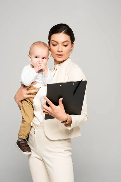 Junge Geschäftsfrau mit kleinen Jungen auf Händen, die mit Klemmbrett arbeiten, isoliert auf grau — Stockfoto