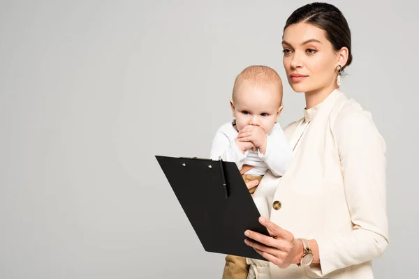 Hermosa mujer de negocios con pequeño bebé en las manos sujetando portapapeles con pluma, aislado en gris - foto de stock