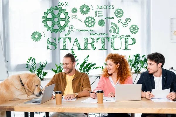 Three friends smiling and looking at cute golden retriever in office with startup illustration — Stock Photo