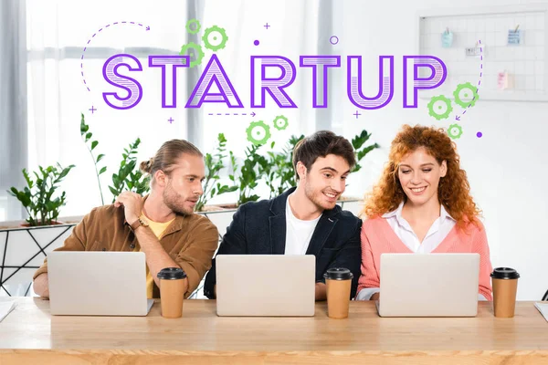 Three smiling friends sitting at table and using laptops in office with startup illustration — Stock Photo