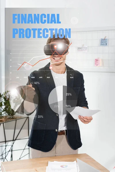 Улыбающийся бизнесмен в рубашке с гарнитурой виртуальной реальности, держащий ручку и бумаги рядом с иллюстрацией финансовой защиты — стоковое фото