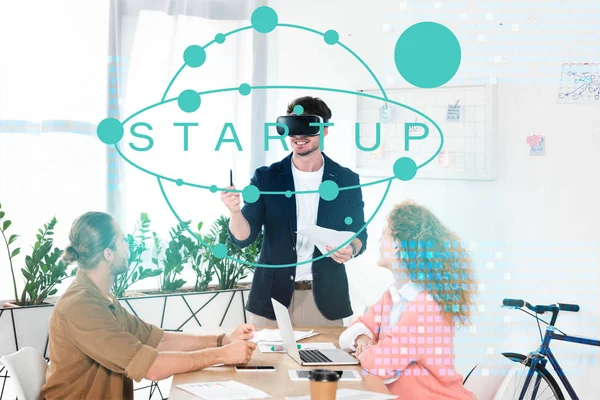 Lächelnder Geschäftsmann mit vr-Headset im Büro in der Nähe von Kollegen und Startup-Illustration — Stockfoto