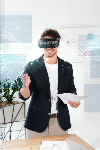 Smiling businessman in shirt with virtual reality headset holding pen and papers near startup illustration — Stock Photo