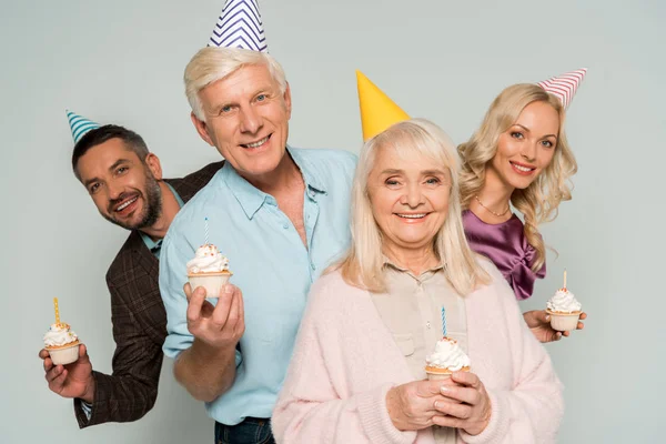 Padres ancianos felices con hija adulta e hijo sosteniendo cupcakes de cumpleaños aislados en gris - foto de stock