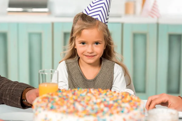 Селективное внимание очаровательного ребенка в кепке для вечеринок, смотрящего в камеру, сидя рядом с праздничным тортом — стоковое фото