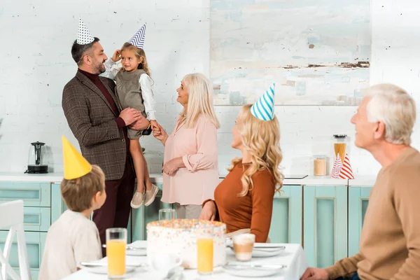 Happy man holding daughter on hands while standing with family near birthday cake on kitchen table — Stock Photo