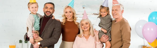 Panoramic shot of happy family posing at camera in party caps — Stock Photo