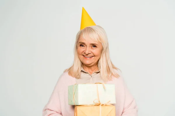 Happy senior woman in party cap holding gift boxes isolated on grey — Stock Photo