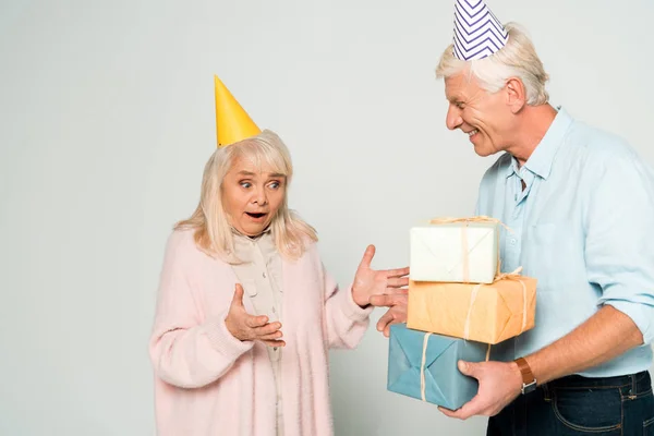 Sonriente hombre mayor presentando cajas de regalo a esposa sorprendida sobre fondo gris - foto de stock