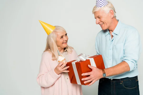 Sonriente hombre mayor presentando caja de regalo a feliz esposa celebración cupcake cumpleaños aislado en gris - foto de stock