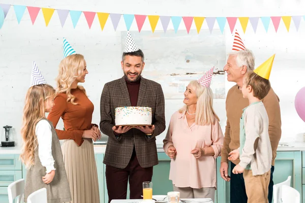 Homme heureux tenant gâteau d'anniversaire tout en se tenant près de la famille joyeuse — Photo de stock