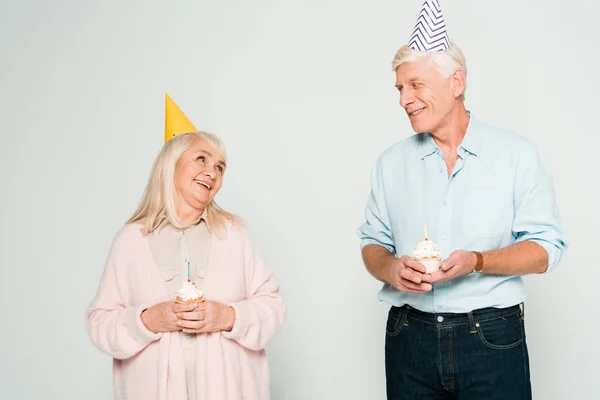 Happy senior husband and wife holding birthday cupcakes and looking at each other isolated on grey — Stock Photo