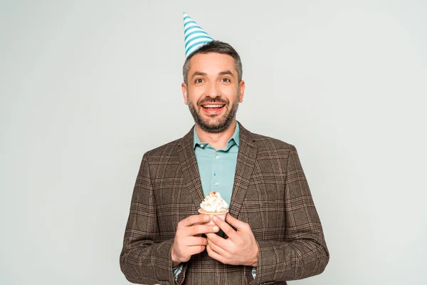 Felice uomo in cappello partito tenendo compleanno cupcake e guardando la fotocamera isolata su grigio — Foto stock