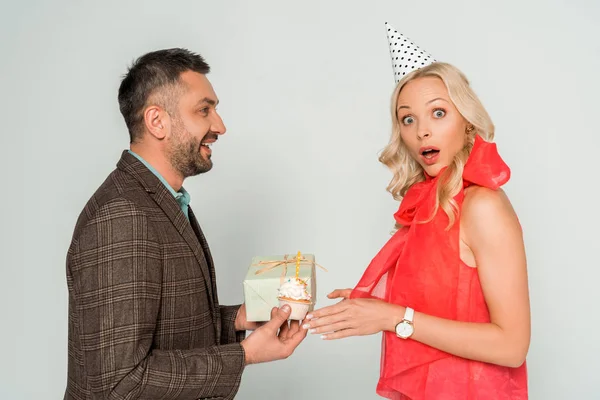 Femme surprise regardant la caméra près du mari présentant boîte cadeau et cupcake d'anniversaire sur fond gris — Photo de stock