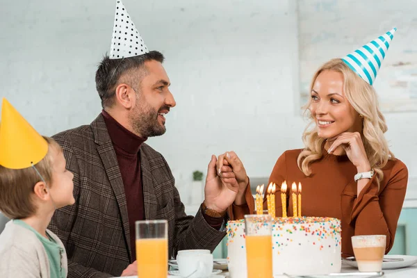 Homme et femme heureux tenant la main tout en étant assis à table avec fils près du gâteau d'anniversaire avec des bougies allumées — Photo de stock