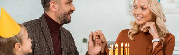 Панорамный снимок счастливого мужчины и женщины, держащихся за руки, сидящих за столом с сыном рядом с праздничным тортом с горящими свечами — стоковое фото