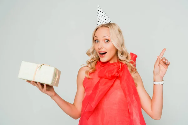 Überraschte Frau blickt in die Kamera, während sie Geschenkschachtel hält und mit dem Finger isoliert auf grau zeigt — Stockfoto