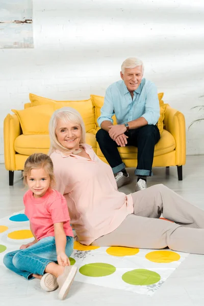 KYIV, UKRAINE - NOVEMBER 21, 2019: senior woman sitting on twister game mat with granddaughter near husband sitting on sofa — Stock Photo