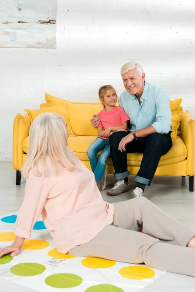 KYIV, UKRAINE - NOVEMBER 21, 2019: senior woman sitting on twister game mat near husband and grandson sitting on sofa — Stock Photo