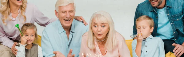 Panoramic shot of senior woman blowing out candles on birthday cake while sitting on sofa near happy family — Stock Photo