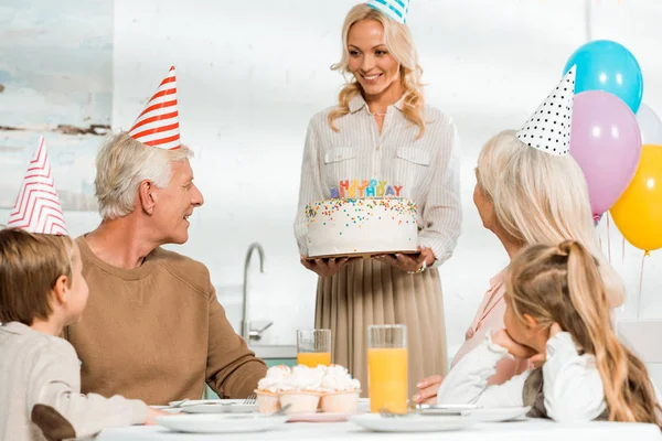 Hermosa mujer presentando pastel de cumpleaños al hombre mayor sentado en la mesa de la cocina cerca de la familia - foto de stock