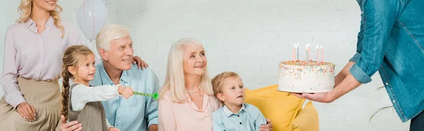 Abgeschnittene Ansicht eines Mannes, der einer Seniorin Geburtstagstorte überreicht, die in der Nähe der Familie auf dem Sofa sitzt, Panoramaaufnahme — Stockfoto