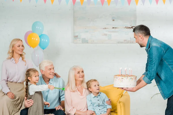 Schöner Mann überreicht Geburtstagstorte an Seniorin, die auf Sofa neben Familie sitzt — Stockfoto