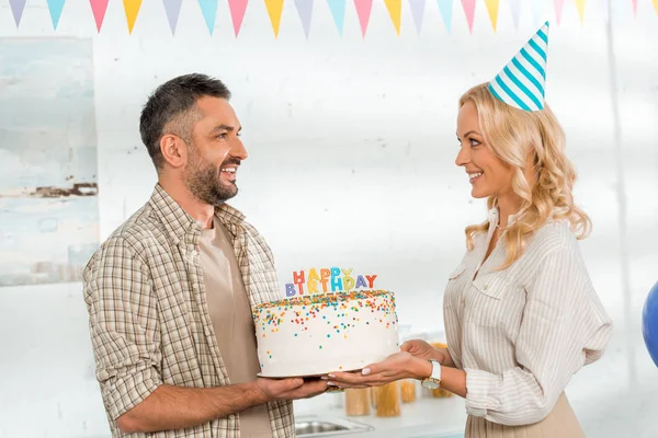 Smiling wife presenting birthday cake with happy birthday candles to husband — Stock Photo
