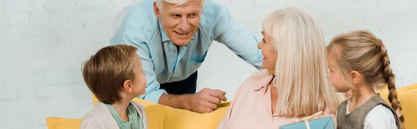 Panoramic shot of senior man talking to happy wife sitting on sofa with grandchildren — Stock Photo