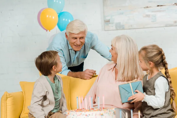 Smiling senior man talking to happy wife sitting on sofa with grandchildren — Stock Photo