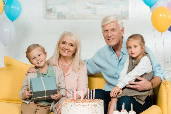 Abuelos felices sentados en el sofá con nietos y sonriendo a la cámara - foto de stock