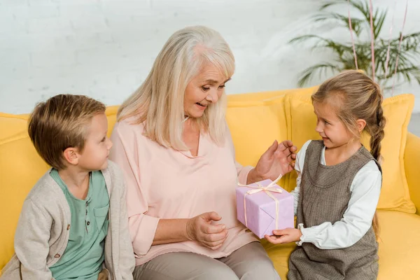 Happy senior woman taking gift box from granddaughter while sitting on yellow sofa with grandson — Stock Photo