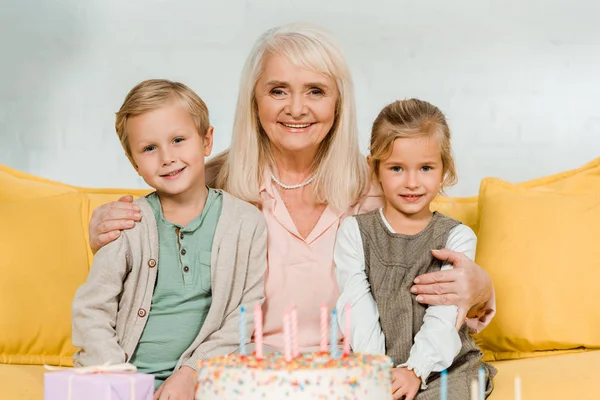 Happy granny embracing grandchildren while sitting near birthday cake and smiling at camera — Stock Photo