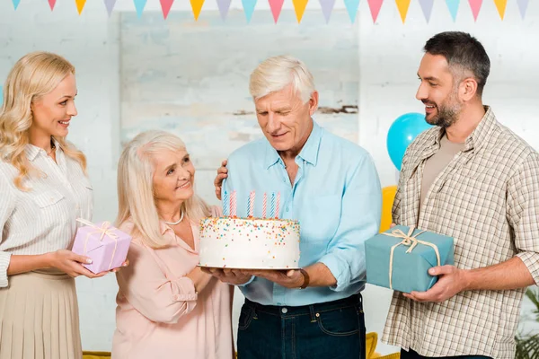 Homem sênior feliz segurando bolo de aniversário enquanto está perto da família — Fotografia de Stock