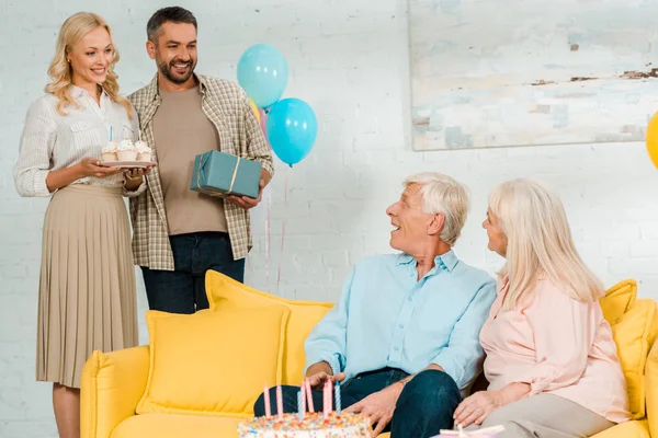 Smiling man and woman holding gift box and birthday cupcakes while standing near happy senior parents — Stock Photo