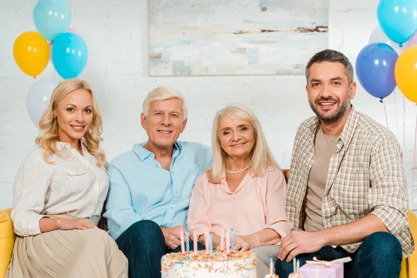 Happy family sitting on yellow sofa near birthday cake and smiling at camera — Stock Photo