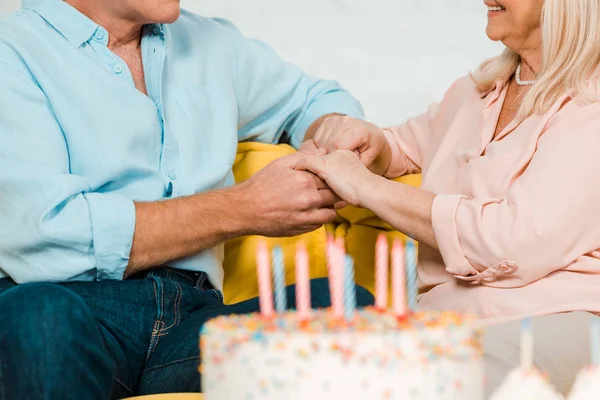 Abgeschnittene Ansicht des älteren Ehepaares, das in der Nähe der Geburtstagstorte sitzt und sich an den Händen hält — Stockfoto